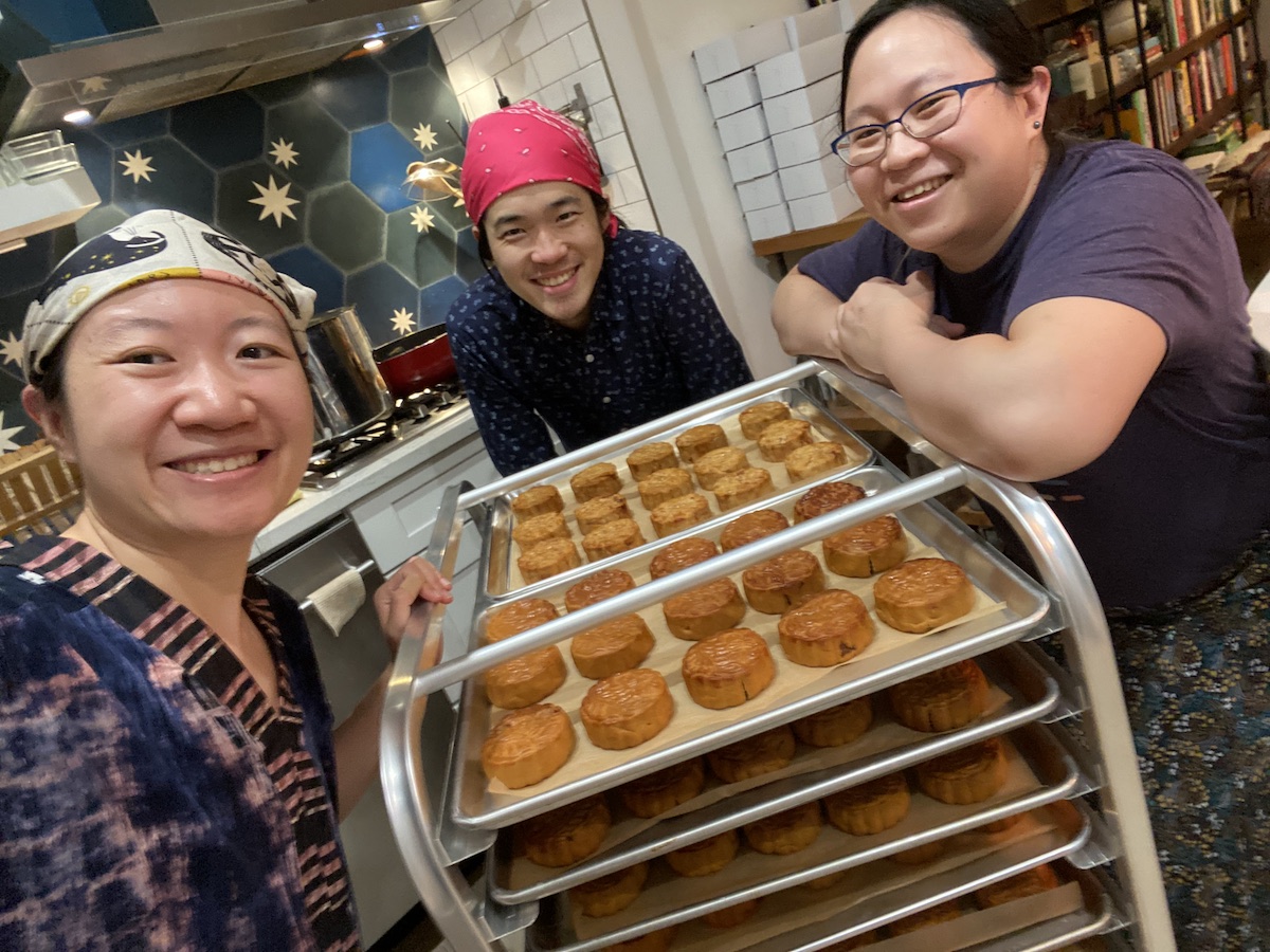 Cordelia, Yang and Tina smiling around a bakery rack full of mooncakes